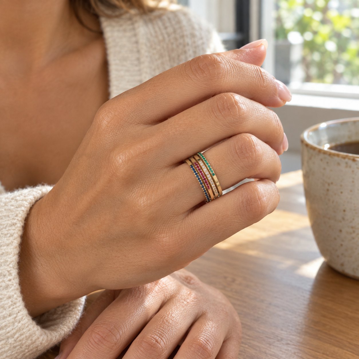 Several Pomence Birthstone Stacking Ring bands laid out together beside a hand wearing a stack of them