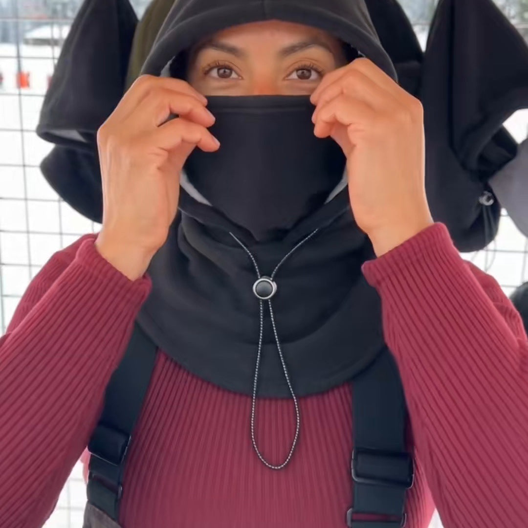 A woman at a snowy ski-hill stall pulls the black face cover up over her nose, the tall collar over her shoulders, real snow and the hood rack behind her.