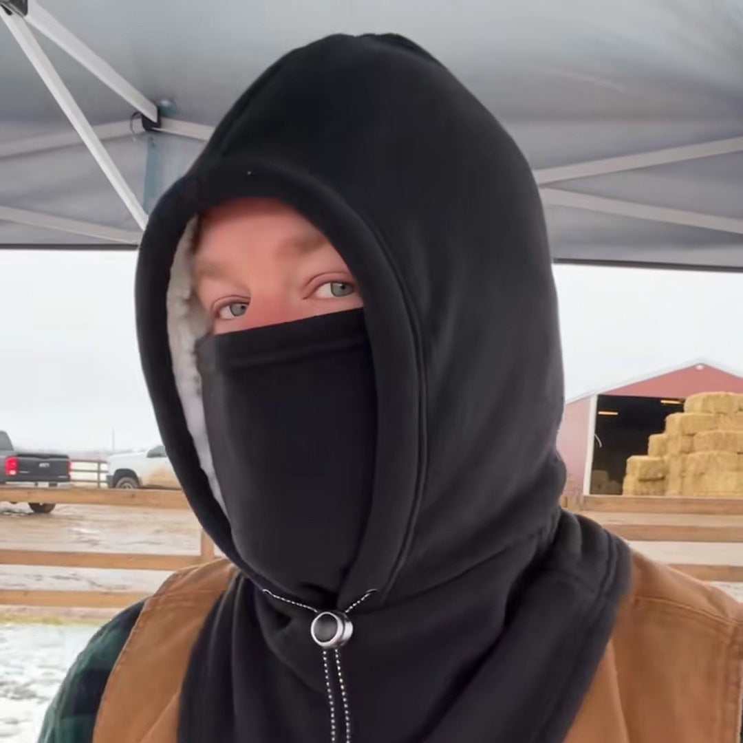 A bearded man at a snowy farm stall with the black hood up and the face cover over his nose, eyes clear, a red barn and hay bales behind him.