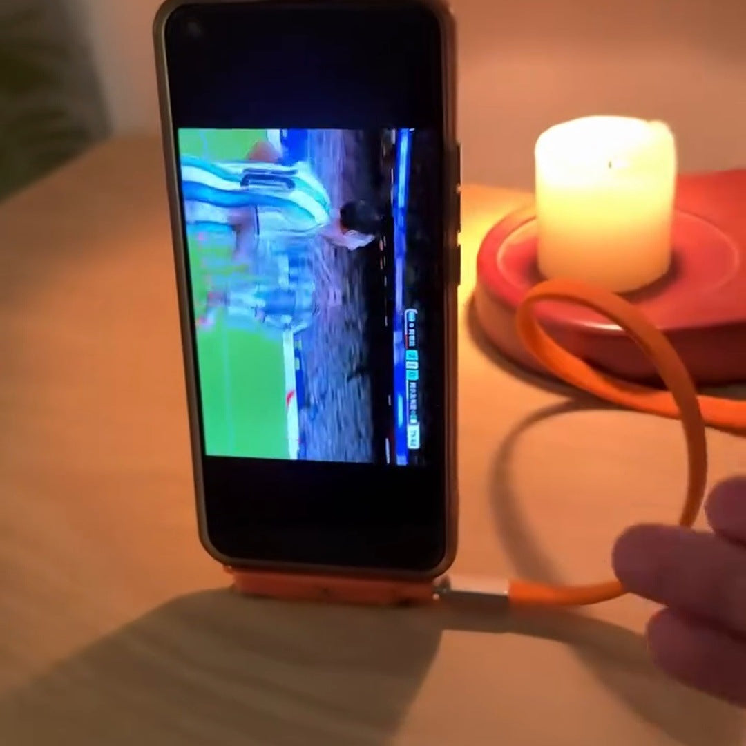 real frame: phone standing upright on the orange plug on a wood desk, soccer on screen, candle behind