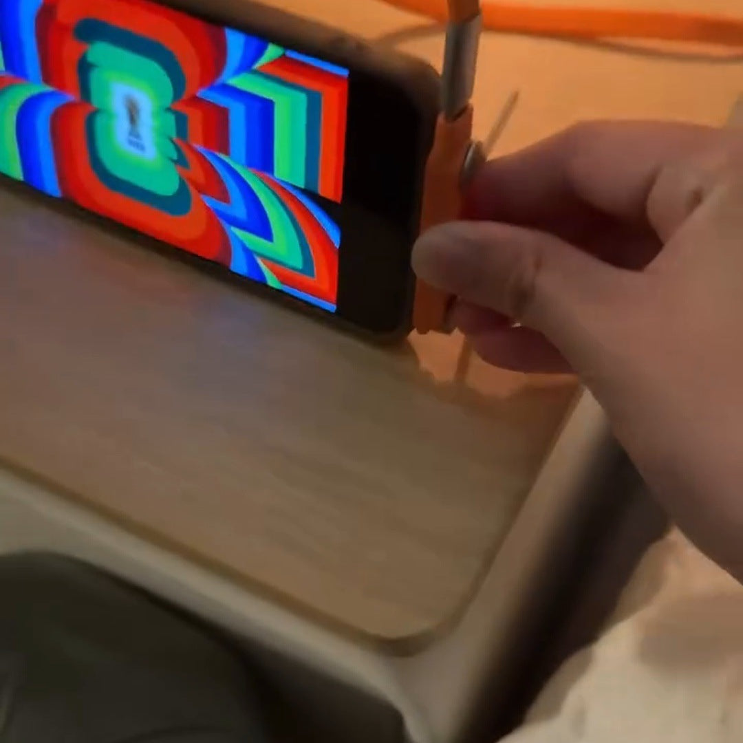 real frame: a phone lying sideways on the orange plug on a wooden desk playing a colourful video, a hand at the plug, the orange cable behind