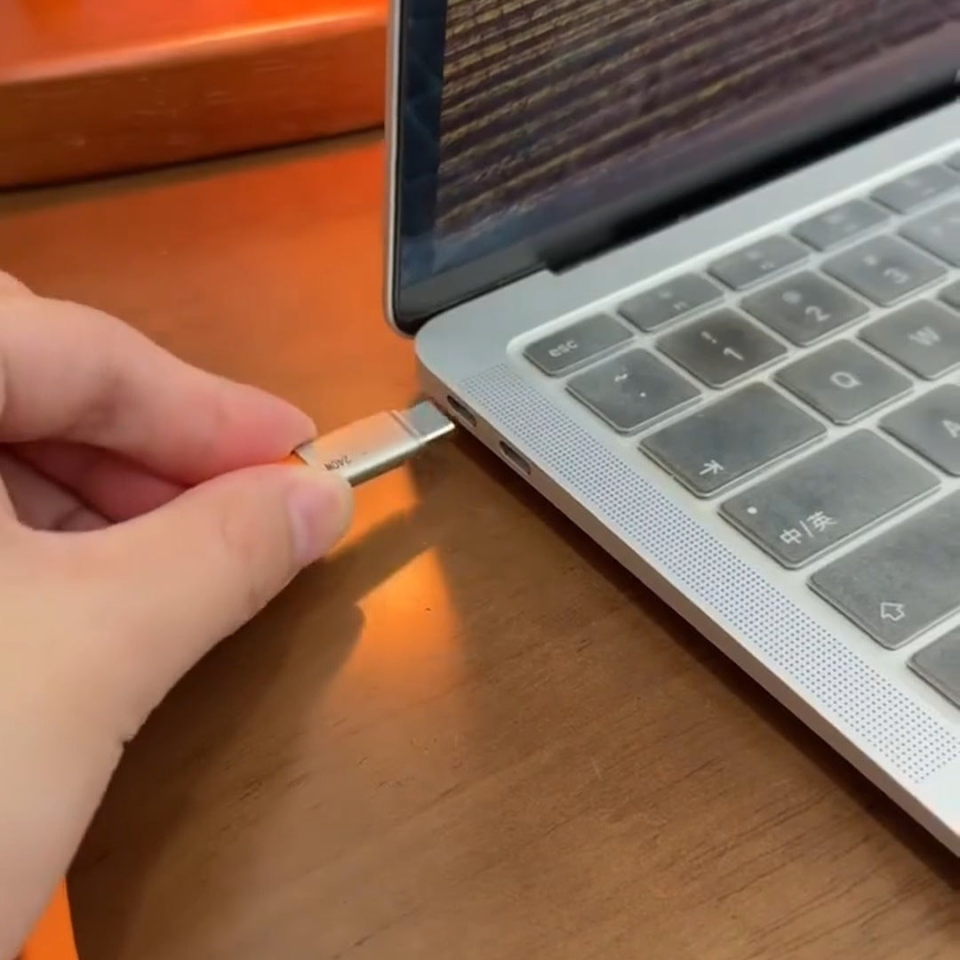 real frame: a hand plugging the flat orange cable into the USB-C port on the side of a silver laptop, the keyboard beside