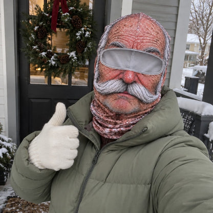 An adult in a sage puffer coat wearing the Grandpa mask on a snowy porch with a wreath.