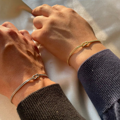 Two bare wrists together, one wearing the Pomence Forever Knot Bracelet in warm 18k-gold tone and one in cool steel-silver tone, both love knots reading