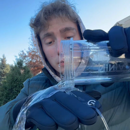 A young man in a green puffer pouring water over the back of his black glove in a fall backyard.