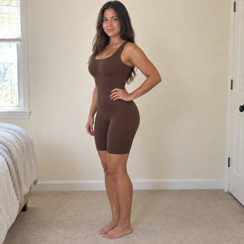 A woman stands side-on in a sunlit bedroom wearing the brown bodysuit, hand on hip, smooth tummy and lifted butt, cream walls set off the brown.