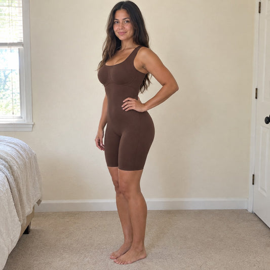 A woman stands side-on in a sunlit bedroom wearing the brown bodysuit, hand on hip, smooth tummy and lifted butt, cream walls set off the brown.
