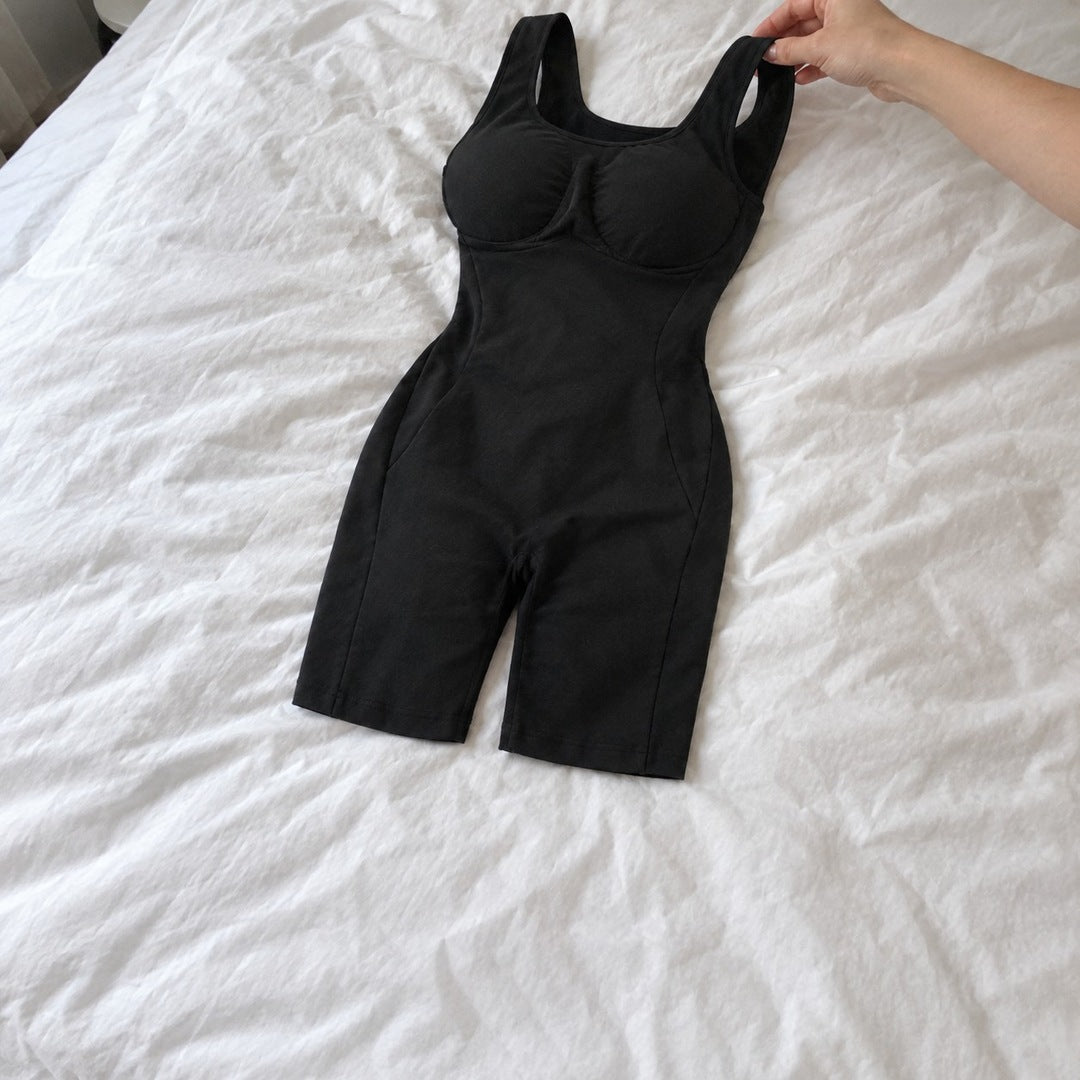 Hands lift one wide shoulder strap of the black bodysuit laid on a white duvet, scoop neck and side panels visible.