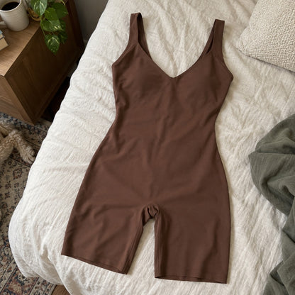 The chocolate boyshort bodysuit lying flat on a cream linen surface, deep plunge V neckline, wide straps relaxed, mid-thigh legs straight