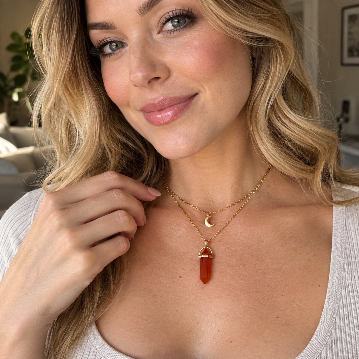 A woman adjusting the gold Pomence Carnelian Attraction Necklace at her throat, the crescent moon charm on the shorter chain above the carnelian point