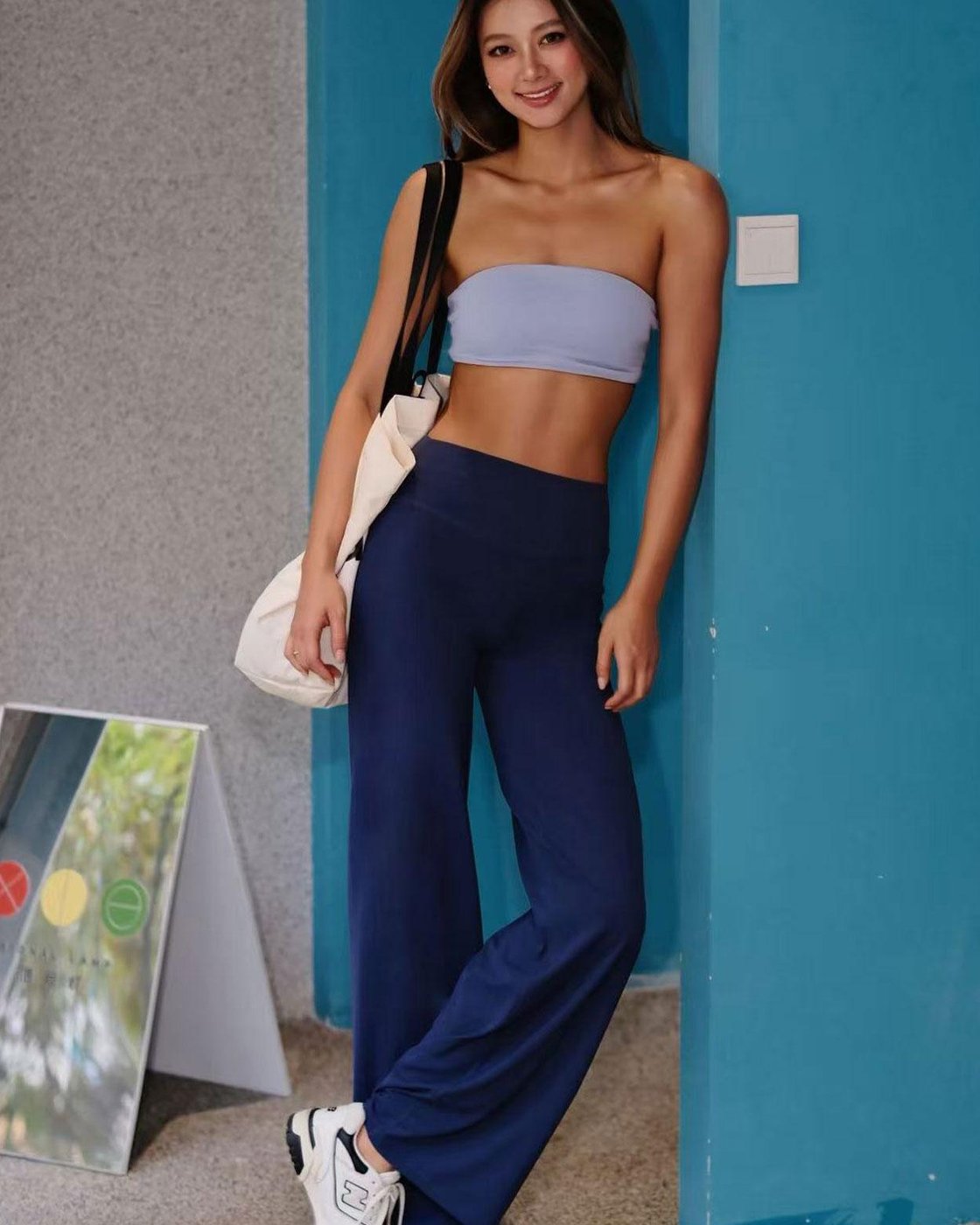 A woman leaning on a blue wall in navy Pomence high-waisted straight leg yoga leggings and white sneakers